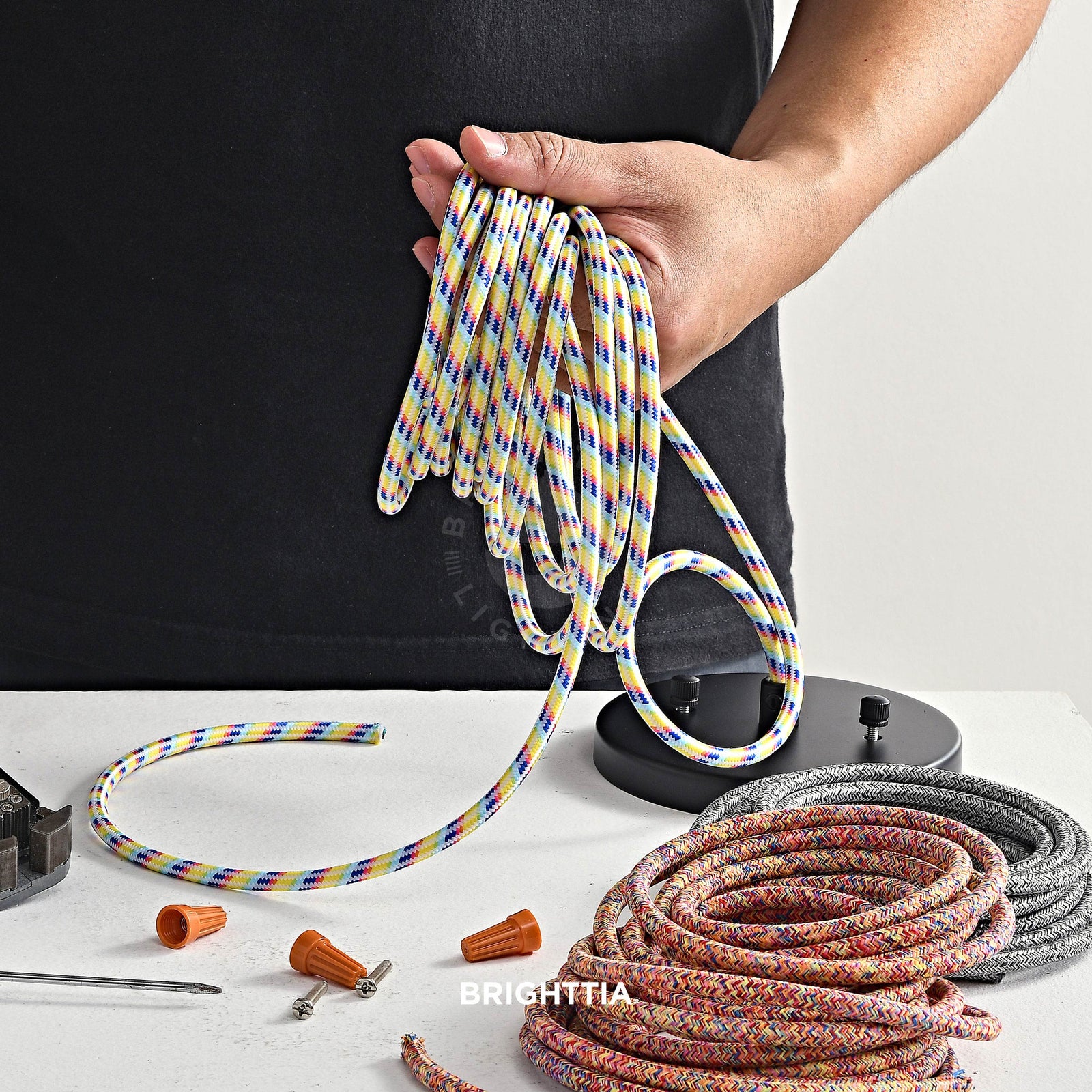 A man holding a roll of rainbow fabric cord on hand making a custom pendant light fixture. A black canopy and two rolls of fabric cord on table by the side.